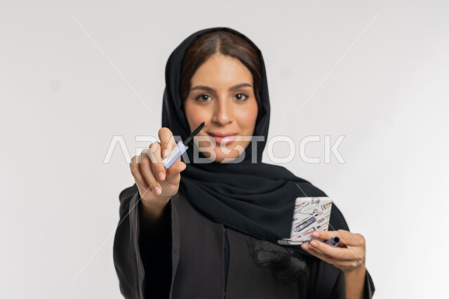 Passion for applying makeup, concept of beautification and beautification, marketing of cosmetics and products, close-up portrait of a veiled Gulf Arab Emirati woman wearing a black abaya holding a mascara brush in her hand, a young Saudi woman looking at the camera with gestures of happiness and pleasure, white background