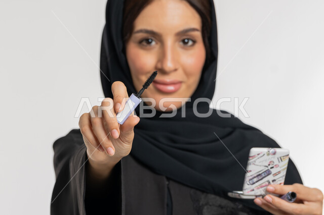 الشغف بوضع المكياج, مفهوم التزين والتجمل, التسويق لمنتجات ومستحضرات التجميل, بورتريه مقرب لامرأة عربية خليجية إماراتية محجبة ترتدي العباءة السوداء تمسك بيدها فرشاة المسكرة, شابة سعودية تنظر إلى الكاميرا بإيماءات السعادة والسرور, خلفية بيضاء