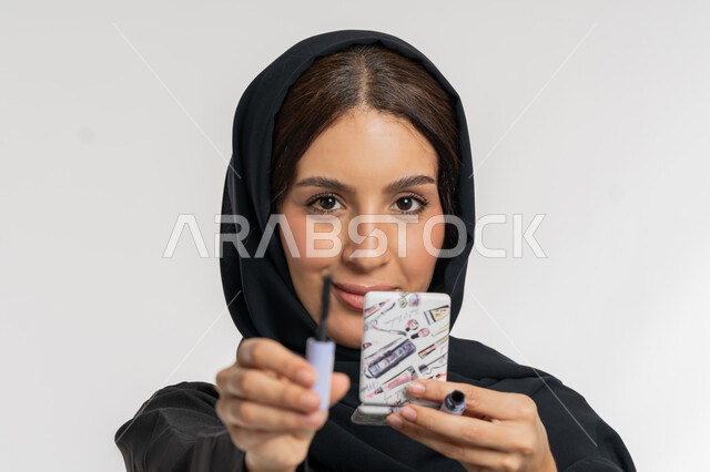 التسويق لمنتجات ومستحضرات التجميل, الشغف بوضع المكياج, مفهوم التزين والتجمل,  بورتريه مقرب لامرأة عربية خليجية إماراتية محجبة ترتدي العباءة السوداء تمسك بيدها فرشاة المسكرة, شابة سعودية تنظر إلى الكاميرا بإيماءات السعادة والسرور, خلفية بيضاء