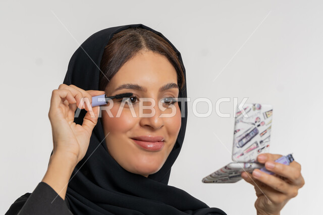 الترويج لمنتجات ومستحضرات التجميل, الاهتمام بالجمال والمظهر الخارجي, مفهوم التزين والتجمل, بورتريه مقرب لامرأة عربية خليجية إماراتية محجبة ترتدي العباءة السوداء تقوم باستخدام المسكرة, شابة سعودية تضع المكياج, خلفية بيضاء