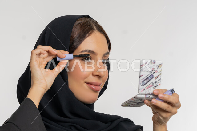 الترويج لمنتجات ومستحضرات التجميل, الاهتمام بالجمال والمظهر الخارجي, مفهوم التزين والتجمل, بورتريه مقرب لامرأة عربية خليجية إماراتية محجبة ترتدي العباءة السوداء تقوم باستخدام المسكرة, شابة سعودية تضع المكياج, خلفية بيضاء
