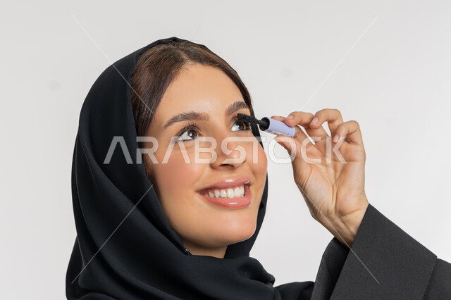 مفهوم التزين والتجمل, الترويج لمنتجات ومستحضرات التجميل, الاهتمام بالجمال والمظهر الخارجي,  بورتريه مقرب لامرأة عربية خليجية إماراتية محجبة ترتدي العباءة السوداء تقوم باستخدام المسكرة, شابة سعودية تضع المكياج, خلفية بيضاء