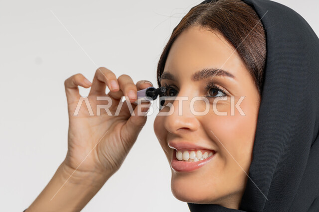 مفهوم التزين والتجمل, الترويج لمنتجات ومستحضرات التجميل, الاهتمام بالجمال والمظهر الخارجي,  بورتريه مقرب لامرأة عربية خليجية إماراتية محجبة ترتدي العباءة السوداء تقوم باستخدام المسكرة, شابة سعودية تضع المكياج, خلفية بيضاء