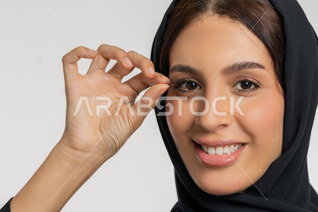 Marketing of cosmetics and products, concept of beautification and beautification, interest in beauty and appearance, close-up portrait of a veiled Emirati Gulf Arab woman wearing a black abaya applying false eyelashes, a young Saudi woman applying makeup, white background