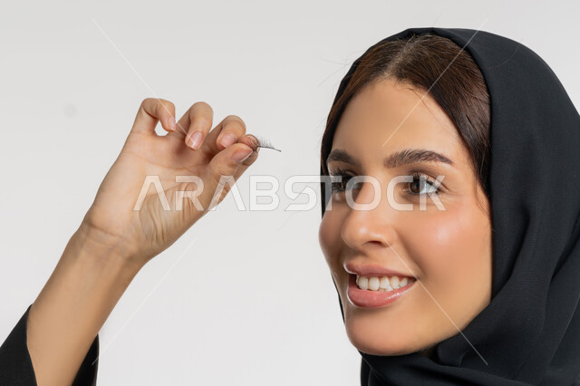 Concern for beauty and appearance, marketing of cosmetics and products, concept of beautification and beautification, close-up portrait of a veiled Emirati Gulf Arab woman wearing a black abaya applying false eyelashes, a young Saudi woman applying makeup, white background