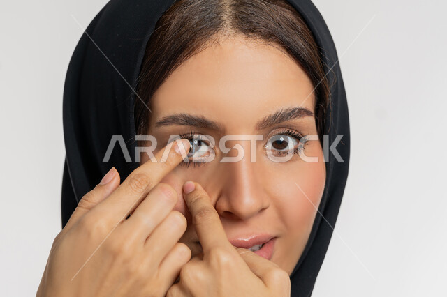 تغير لون العين بواسطة العدسة التجميلية، مفهوم تصحيح النظر وطب العيون، بورتريه مقرب لامرأة عربية خليجية اماراتية ترتدي الحجاب تقوم بوضع العدسات اللاصقة في عينها، شغف التزيين والتجميل للنساء، شابة سعودية تهتم بالاناقة والمظهر الخارجي، خلفية بيضاء