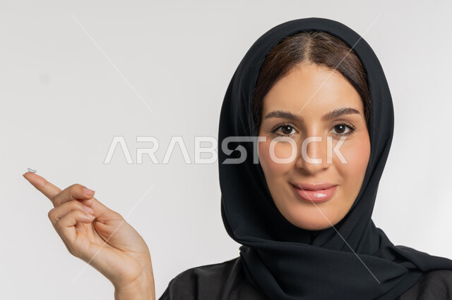 Passion for decoration and beautification for women, a smiling young Saudi woman interested in elegance and external appearance, the concept of vision correction and ophthalmology, changing eye color with cosmetic lenses, a close-up portrait of an Arab Gulf Emirati woman wearing a hijab putting a contact lens on her finger, white background