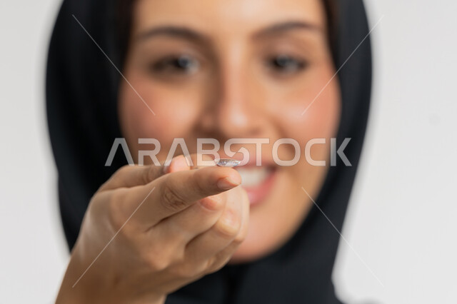 شغف التزيين والتجميل للنساء، شابة سعودية مبتسمة تهتم بالاناقة والمظهر الخارجي، مفهوم تصحيح النظر وطب العيون، تغير لون العين بواسطة العدسات التجميلية، بورتريه مقرب لامرأة عربية خليجية اماراتية ترتدي الحجاب تضع العدسة اللاصقة على اصبعها، خلفية بيضاء