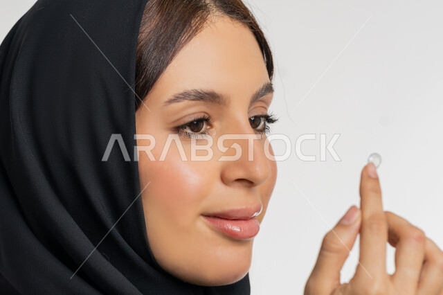 مفهوم تصحيح النظر وطب العيون، تغير لون العين بواسطة العدسة التجميلية، بورتريه مقرب لامرأة عربية خليجية اماراتية ترتدي الحجاب تقوم بوضع العدسات اللاصقة في عينها، شغف التزيين والتجميل للنساء، شابة سعودية تهتم بالاناقة والمظهر الخارجي، خلفية بيضاء