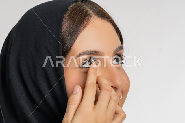 Changing eye color with cosmetic lenses, vision correction and ophthalmology concept, close-up portrait of an Arab Gulf Emirati woman wearing hijab putting contact lenses in her eyes, passion for decoration and beautification for women, a young Saudi woman interested in elegance and external appearance, white background
