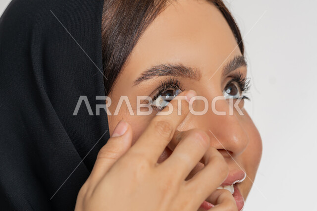 مفهوم تصحيح النظر وطب العيون، تغير لون العين بواسطة العدسة التجميلية، بورتريه مقرب لامرأة عربية خليجية اماراتية ترتدي الحجاب تقوم بوضع العدسات اللاصقة في عينها، شغف التزيين والتجميل للنساء، شابة سعودية تهتم بالاناقة والمظهر الخارجي، خلفية بيضاء