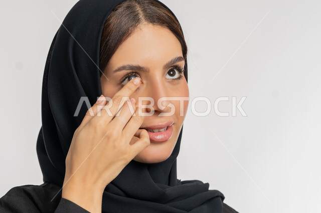 تغير لون العين بواسطة العدسة التجميلية، مفهوم تصحيح النظر وطب العيون، بورتريه مقرب لامرأة عربية خليجية اماراتية ترتدي الحجاب تقوم بوضع العدسات اللاصقة في عينها، شغف التزيين والتجميل للنساء، شابة سعودية تهتم بالاناقة والمظهر الخارجي، خلفية بيضاء
