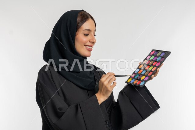 استخدام المستحضرات التجميلية, الترويج لمنتجات و مساحيق التجميل, الاهتمام بالمظهر الخارجي, مفهوم العناية بالجمال والبشرة, بورتريه مقرب لامرأة عربية خليجية إماراتية محجبة ترتدي العباءة السوداء تستعمل فرشاة ظل العيون, شابة سعودية تقوم بوضع المكياج, خلفية بيضاء