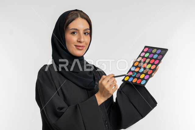 الاهتمام بالمظهر الخارجي, استخدام المستحضرات التجميلية, الترويج لمنتجات و مساحيق التجميل, مفهوم العناية بالجمال والبشرة, بورتريه مقرب لامرأة عربية خليجية إماراتية محجبة ترتدي العباءة السوداء تستعمل فرشاة ظل العيون, شابة سعودية تقوم بوضع المكياج, خلفية بيضاء