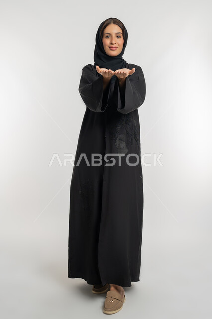 النظر إلى الكاميرا بايماءات السعادة والسرور، بورتريه لامرأة عربية خليجية سعودية محجبة ترتدي عباءة سوداء تفتح يديها لعرض شيء ما، شابة اماراتية تقوم بحمل ورفع عنصر ما على الكفين، صورة الجسم بالطول الكامل، خلفية بيضاء