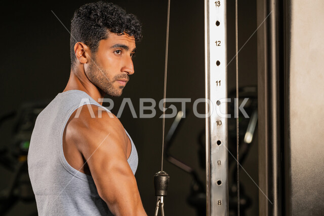استخدام الأجهزة الرياضية، صورة مقربة من الجانب لشاب عربي خليجي إماراتي يرتدي الزي الرياضي يقوم بممارسة تمارين رفع الأوزان، رياضة كمال الأجسام، أسلوب حياة صحي، الاهتمام بالصحة واللياقة الجسدية، إيماءات القوة والثقة، رجل سعودي يمارس أنشطة لتقوية العضلات