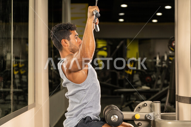 رجل سعودي يمارس أنشطة لتقوية العضلات، رياضة كمال الأجسام، صورة من الجانب لشاب عربي خليجي إماراتي يرتدي الزي الرياضي يقوم بممارسة تمارين رفع الأوزان، استخدام الأجهزة الرياضية، الاهتمام بالصحة واللياقة الجسدية، إيماءات القوة والثقة، بناء الكتلة العضلية للجسم