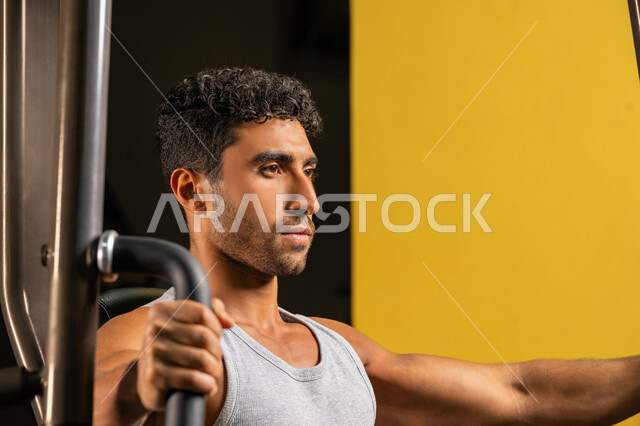 إيماءات القوة والثقة، أسلوب حياة صحي، استخدام الأجهزة الرياضية، حرق الدهون وتقليل الوزن، صورة مقربة لشاب عربي خليجي إماراتي يرتدي الزي الرياضي يقوم بممارسة تمارين رفع الأوزان، رجل سعودي يمارس أنشطة لتقوية العضلات، الاهتمام بالصحة واللياقة، رياضة كمال الأجسام