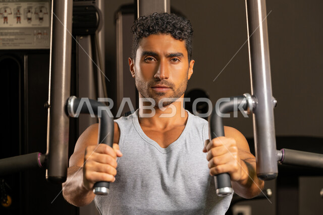 استخدام الأجهزة الرياضية، حرق الدهون وتقليل الوزن، صورة مقربة لشاب عربي خليجي إماراتي يرتدي الزي الرياضي يقوم بممارسة تمارين رفع الأوزان، أسلوب حياة صحي، رجل سعودي يمارس أنشطة لتقوية العضلات، إيماءات القوة والثقة، الاهتمام بالصحة واللياقة، رياضة كمال الأجسام