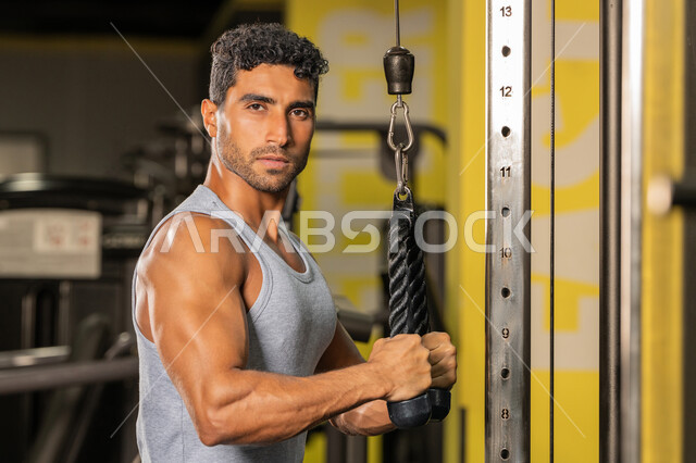 إيماءات القوة والثقة، رياضة كمال الأجسام، أسلوب حياة صحي، الاهتمام بالصحة واللياقة الجسدية، استخدام الأجهزة الرياضية، صورة مقربة من الجانب لشاب عربي خليجي إماراتي يرتدي الزي الرياضي يقوم بممارسة تمارين رفع الأوزان، رجل سعودي يمارس أنشطة لتقوية العضلات