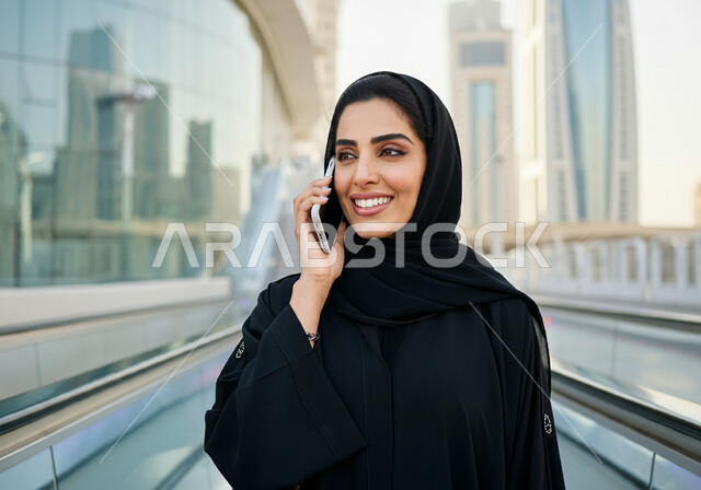 إيماءات السعاده والرضا بالخدمات الحديثه, اجراء مكالمه هاتفيه عبر الجوال, إمرأه عربيه سعوديه خليجيه مبتسمه  ترتدي الحجاب و العباءه السوداء تقف في محطة قطار الرياض, تقدم وسائل المواصلات  في المملكه , التنقل السريع والآمن عبر الميترو في السعوديه , تيسير الحركه المروريه للشعب السعودي
