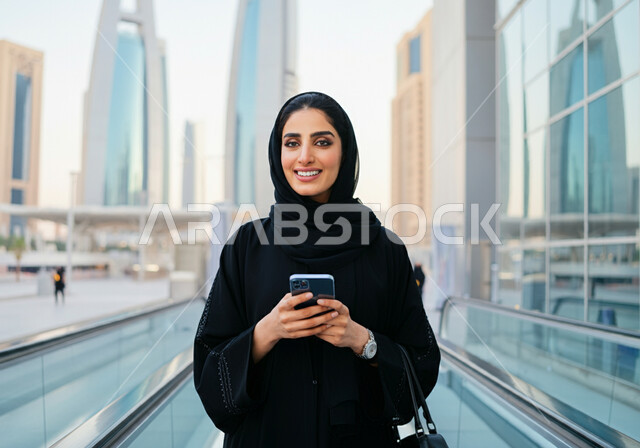 Using a mobile phone, a smiling Saudi Arabian Gulf woman wearing a hijab and a black abaya standing at Riyadh train station, gestures of happiness and satisfaction with modern services, the progress of means of transportation in the Kingdom, fast and safe movement via the metro in Saudi Arabia, facilitating traffic movement for the Saudi people