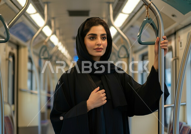 الامساك بالعامود الحديدي داخل القطار للحفاظ على السلامه, إمرأه عربيه سعوديه خليجيه مبتسمه  ترتدي الحجاب و العباءه السوداء تقف في مترو الرياض,  إيماءات الطمانينه والرضا بالخدمات الحديثه ,تقدم وسائل المواصلات  في المملكه , التنقل السريع والآمن عبر الميترو في السعوديه , تيسير الحركه المروريه للشعب السعودي