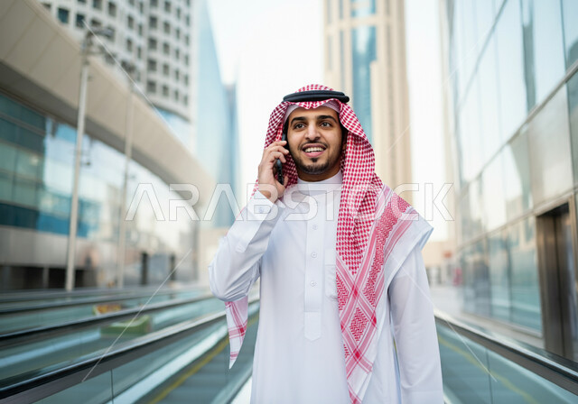 Making a phone call via mobile, a smiling Saudi Arabian Gulf man wearing a thobe and a shemagh standing at Riyadh train station, gestures of happiness and satisfaction with modern services, progress and development of means of transportation in the Kingdom, fast and safe transportation via the metro in Saudi Arabia, facilitating traffic movement for the Saudi people