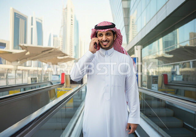 Making a phone call via mobile, a smiling Saudi Arabian Gulf man wearing a thobe and a shemagh standing at Riyadh train station, gestures of happiness and satisfaction with modern services, progress and development of means of transportation in the Kingdom, fast and safe transportation via the metro in Saudi Arabia, facilitating traffic movement for the Saudi people