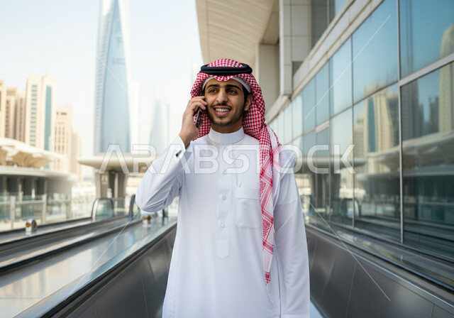 Making a phone call via mobile, a smiling Saudi Arabian Gulf man wearing a thobe and a shemagh standing at Riyadh train station, gestures of happiness and satisfaction with modern services, progress and development of means of transportation in the Kingdom, fast and safe transportation via the metro in Saudi Arabia, facilitating traffic movement for the Saudi people