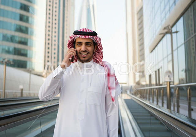 Using a mobile phone, a smiling Saudi Arabian Gulf man wearing a thobe and a shemagh standing at Riyadh train station, gestures of happiness and satisfaction with modern services, progress and development of means of transportation in the Kingdom, fast and safe transportation via the metro in Saudi Arabia, facilitating traffic movement for the Saudi people