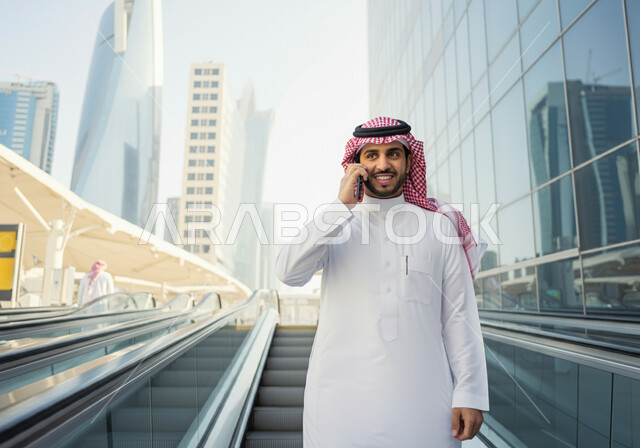 استخدام الهاتف المحمول, رجل عربي سعودي خليجي مبتسم  يرتدي الثوب والشماغ يقف في محطة قطار الرياض,  إيماءات السعاده والرضا بالخدمات الحديثه, تقدم وتطور وسائل المواصلات  في المملكه, التنقل السريع والآمن عبر الميترو في السعوديه, تيسير الحركه المروريه للشعب السعودي