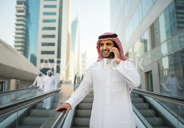 استخدام الهاتف المحمول, رجل عربي سعودي خليجي مبتسم  يرتدي الثوب والشماغ يقف في محطة قطار الرياض,  إيماءات السعاده والرضا بالخدمات الحديثه, تقدم وتطور وسائل المواصلات  في المملكه, التنقل السريع والآمن عبر الميترو في السعوديه, تيسير الحركه المروريه للشعب السعودي