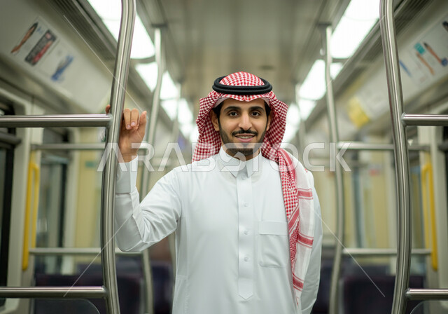 وسائل المواصلات في المملكة, تيسير الحركة المرورية للشعب السعودي, الإمساك بالعامود الحديدي للحفاظ على السلامة, صورة مقربة لرجل عربي خليجي سعودي مبتسم يرتدي الثوب التقليدي والشماغ  يقف في مترو الرياض, النظر إلى الكاميرا بإيماءات السعادة والرضا بالخدمات الحديثة