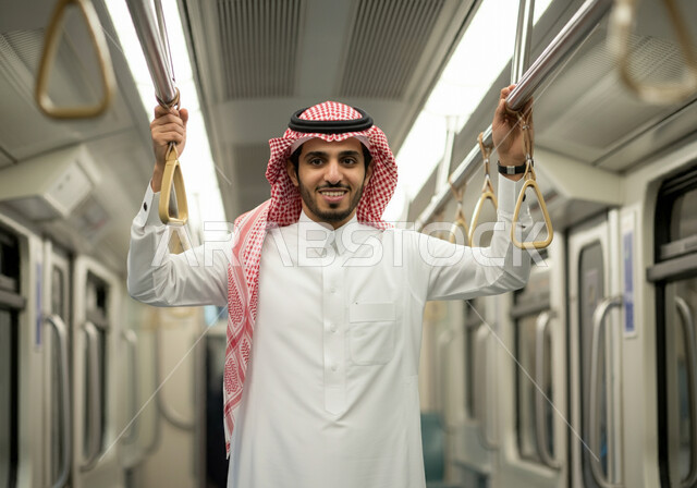 مفهوم تطور وسائل المواصلات في المملكة, تيسير الحركة المرورية للشعب السعودي, الإمساك بالعامود الحديدي للحفاظ على السلامة, رجل عربي خليجي سعودي مبتسم يرتدي الثوب التقليدي والشماغ  يقف في مترو الرياض, النظر إلى الكاميرا بإيماءات السعادة والرضا بالخدمات الحديثة