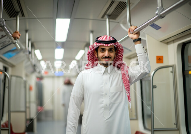النظر إلى الكاميرا بإيماءات السعادة والرضا بالخدمات الحديثة,  وسائل المواصلات في المملكة, تيسير الحركة المرورية للشعب السعودي, الإمساك بالعامود الحديدي للحفاظ على السلامة, صورة مقربة لرجل عربي خليجي سعودي مبتسم يرتدي الثوب التقليدي والشماغ  يقف في مترو الرياض