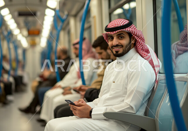 النظر إلى الكاميرا بإيماءات السعادة والسرور, استخدام الأجهزة التقنية الحديثة, توفير خدمات التنقل السريع والآمن, تطور وسائل النقل والمواصلات في المملكة, رجل عربي خليجي سعودي يرتدي الثوب التقليدي والشماغ يجلس داخل مترو الرياض ويستعمل الهاتف المحمول