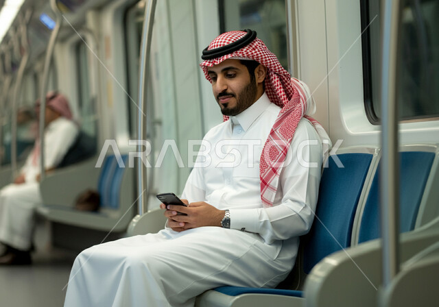 Using modern technical devices, providing fast and safe transportation services, development of transportation and communication methods in the Kingdom, a Saudi Gulf Arab man wearing the traditional dress and shemagh sitting inside the Riyadh Metro and using a mobile phone