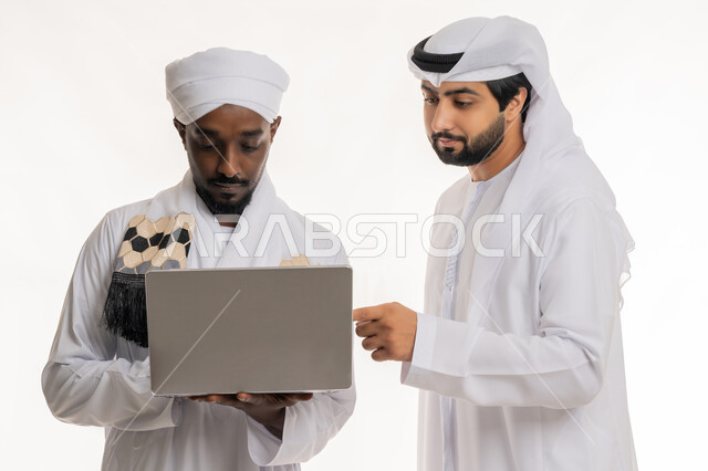 إنجاز مهام العمل باستخدام الأجهزة التقنية الحديثة والمتطورة, تبادل الخبرات والمهارات, مناقشة الخطط والمشاريع, بورتريه مقرب لشاب عربي خليجي إماراتي يرتدي الكندورة والغترة البيضاء يعمل مع رجل سوداني, خلفية بيضاء