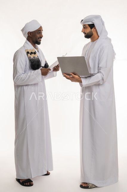 استخدام الأجهزة التقنية الحديثة والمتطورة, التعاون في إنجاز مهام العمل, تبادل الخبرات والمهارات, مناقشة الخطط والمشاريع, بورتريه لشاب عربي خليجي إماراتي يرتدي الكندورة والغترة البيضاء يعمل مع رجل سوداني, الجسم بالطول الكامل, خلفية بيضاء