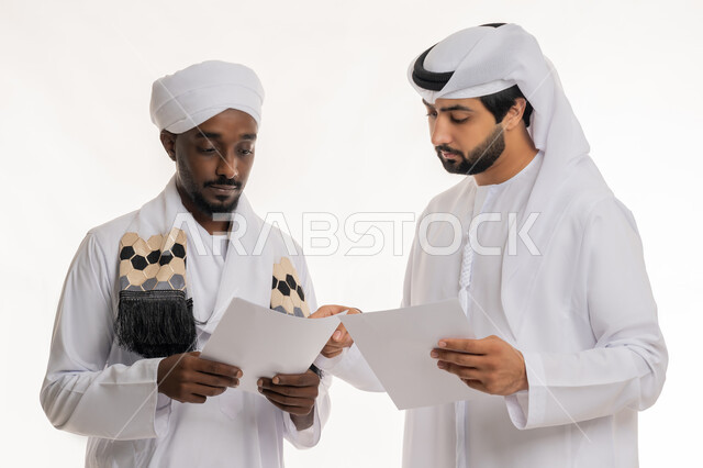 إعداد التقارير الخاصة بالعمل, التدقيق في قراءة الملاحظات, بورتريه مقرب لشاب عربي خليجي إماراتي يرتدي الكندورة والغترة البيضاء يعمل مع رجل سوداني يرتدي الجلابية والعمامة ينظران إلى أوراق بيضاء, مراجعة ومتابعة الخطط والمشاريع بإيماءات التركيز والاندماج, تبادل الخبرات والمهارات, خلفية بيضاء
