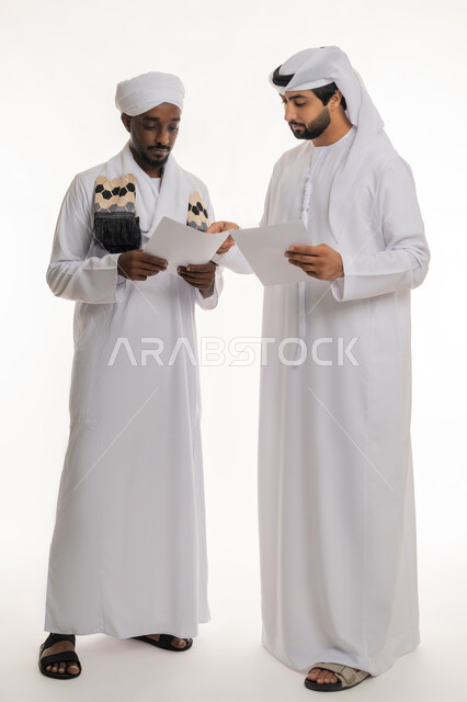 مراجعة ومتابعة الخطط والمشاريع بإيماءات التركيز والاندماج, إعداد التقارير الخاصة بالعمل, بورتريه مقرب لشاب عربي خليجي إماراتي يرتدي الكندورة والغترة البيضاء يعمل مع رجل سوداني يرتدي الجلابية والعمامة ينظران إلى أوراق بيضاء, الجسم بالطول الكامل, خلفية بيضاء