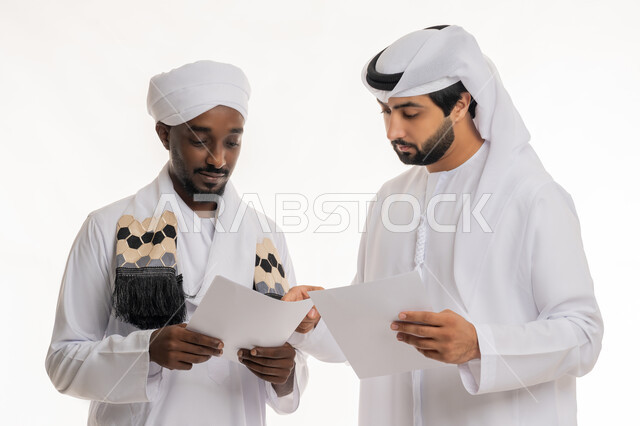 التدقيق في قراءة الملاحظات, إعداد التقارير الخاصة بالعمل, بورتريه مقرب لشاب عربي خليجي إماراتي يرتدي الكندورة والغترة البيضاء يعمل مع رجل سوداني يرتدي الجلابية والعمامة ينظران إلى أوراق بيضاء, مراجعة ومتابعة الخطط والمشاريع بإيماءات التركيز والاندماج, تبادل الخبرات والمهارات, خلفية بيضاء