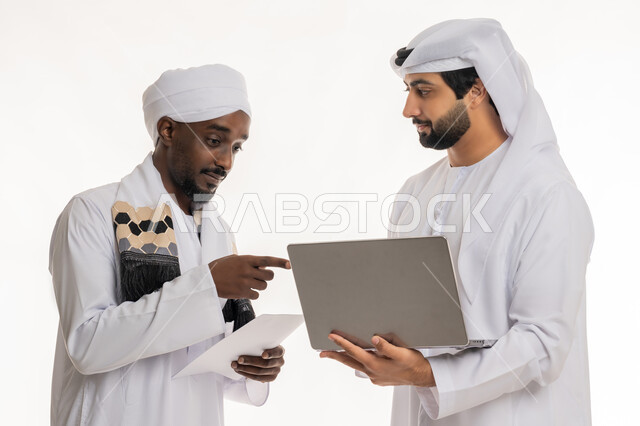 الإشارة بإصبع السبابة إلى شيء ما, تبادل الخبرات والمهارات, مناقشة الخطط والمشاريع, التعاون في إنجاز مهام العمل, استخدام الأجهزة التقنية الحديثة والمتطورة, بورتريه مقرب لشاب عربي خليجي إماراتي يرتدي الكندورة والغترة البيضاء يعمل مع رجل سوداني, خلفية بيضاء