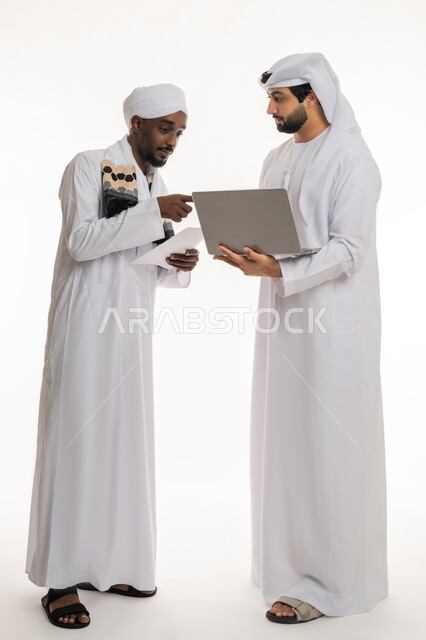 تبادل الخبرات والمهارات, الإشارة بإصبع السبابة إلى شيء ما, التعاون في إنجاز مهام العمل, استخدام الأجهزة التقنية الحديثة والمتطورة, بورتريه لشاب عربي خليجي إماراتي يرتدي الكندورة والغترة البيضاء يعمل مع رجل سوداني, الجسم بالطول الكامل, خلفية بيضاء