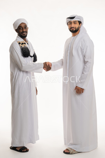 مفهوم التعارف والصداقات, بناء علاقات اجتماعية جيدة, إلقاء التحية والسلام, النظر إلى الكاميرا بإيماءات السعادة والسرور, بورتريه مقرب لشاب عربي خليجي إماراتي يرتدي الكندورة والغترة البيضاء يقوم بمصافحة رجل سودني, الجسم بالطول الكامل, خلفية بيضاء
