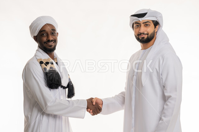 بناء علاقات اجتماعية جيدة, إلقاء التحية والسلام, مفهوم التعارف والصداقات, النظر إلى الكاميرا بإيماءات السعادة والسرور, بورتريه مقرب لشاب عربي خليجي إماراتي يرتدي الكندورة والغترة البيضاء يقوم بمصافحة رجل سودني, خلفية بيضاء