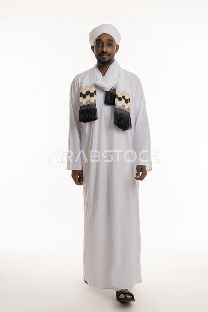 النظر إلى الكاميرا بإيماءات السعادة والسرور, الذهاب إلى مكان ما, مفهوم الأناقة والرجولة, الاهتمام بالمظهر الخارجي, بورتريه لرجل عربي سوداني مبتسم يرتدي الجلابية والعمامة السودانية يقوم بالمشي, الجسم بالطول الكامل, خلفية بيضاء