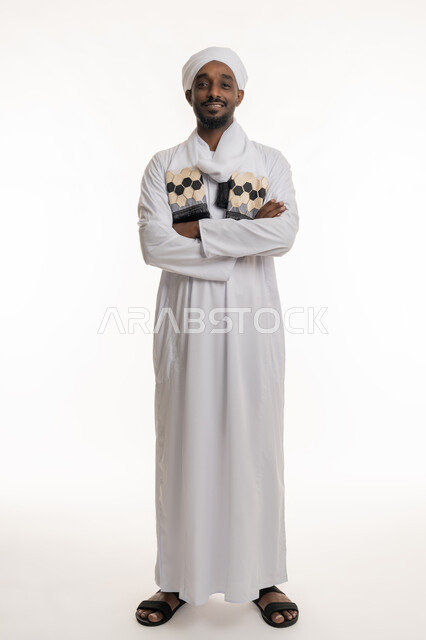 الاهتمام بالمظهر الخارجي, النظر إلى الكاميرا بإيماءات السعادة والسرور, مفهوم الأناقة والرجولة, بورتريه لرجل عربي سوداني مبتسم يرتدي الجلابية والعمامة السودانية يقف بأيدي متقاطعة بتعابير الثقة بالنفس, خلفية بيضاء