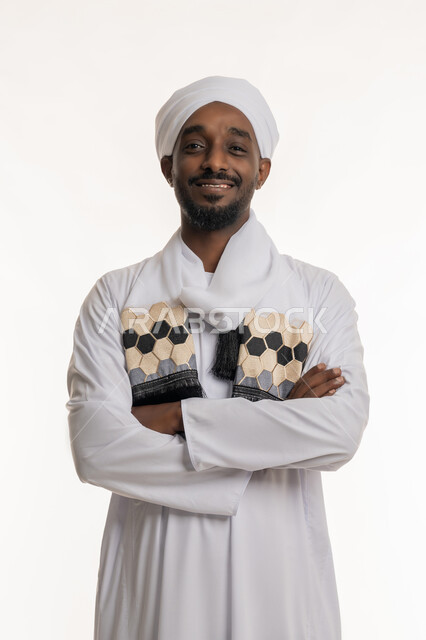 النظر إلى الكاميرا بإيماءات السعادة والسرور, الاهتمام بالمظهر الخارجي, مفهوم الأناقة والرجولة, بورتريه لرجل عربي سوداني مبتسم يرتدي الجلابية والعمامة السودانية يقف بأيدي متقاطعة بتعابير الثقة بالنفس, خلفية بيضاء